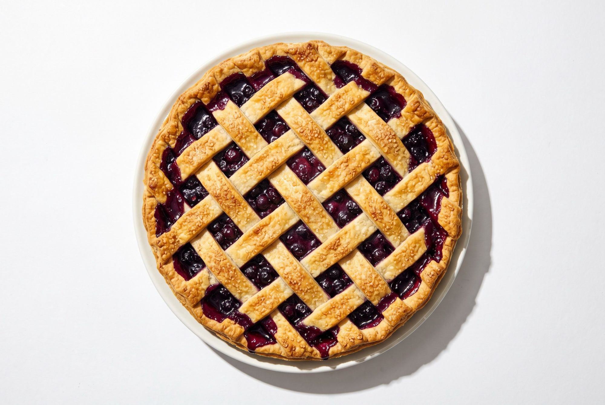 A whole blueberry pie with a woven lattice crust and dark filling bubbling through, seen from above on a white backdrop