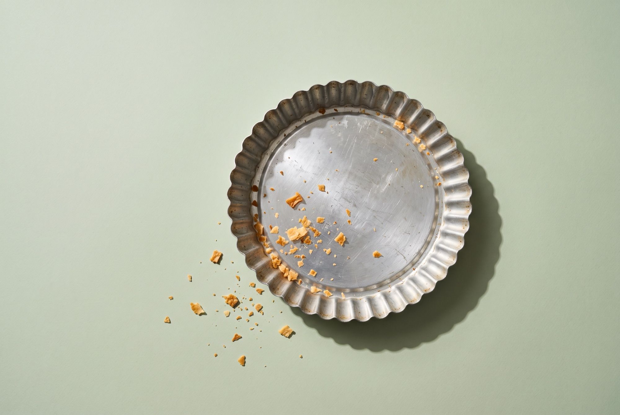 An empty fluted metal pie tin with a few scattered pastry crumbs on a pale green backdrop