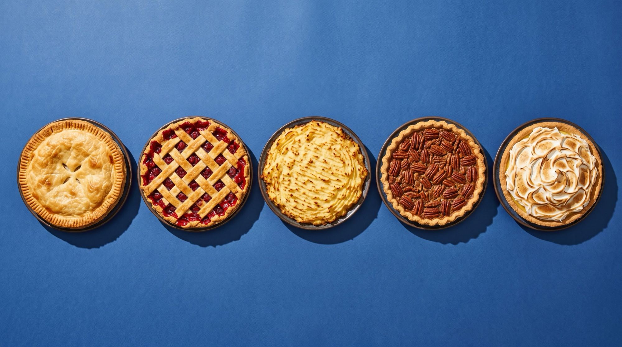 Five different whole pies in a row on a cobalt blue backdrop, seen from above: double-crust, cherry lattice, mash-topped, pecan and meringue