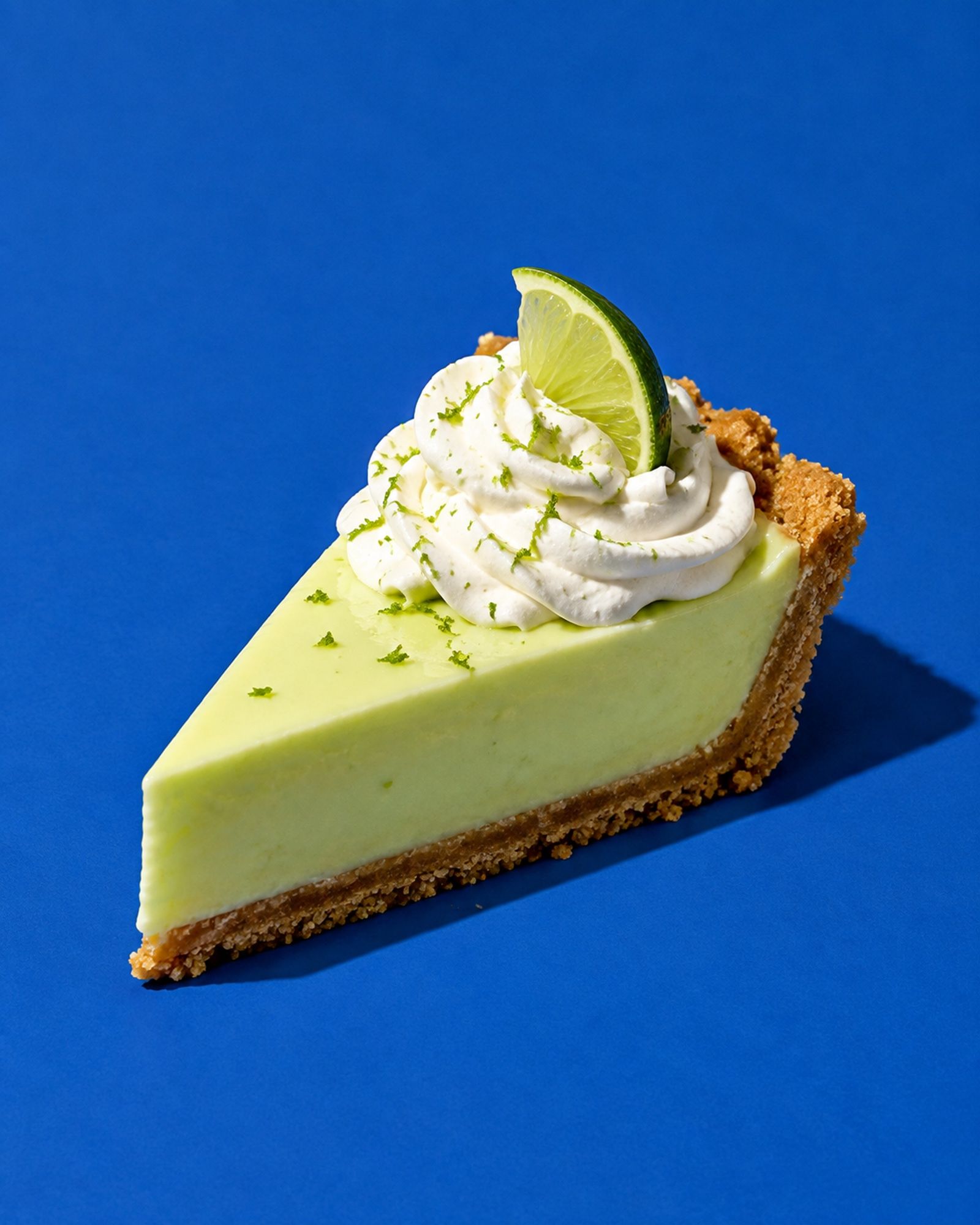 A slice of key lime pie with whipped cream and a lime wedge on a cobalt blue backdrop