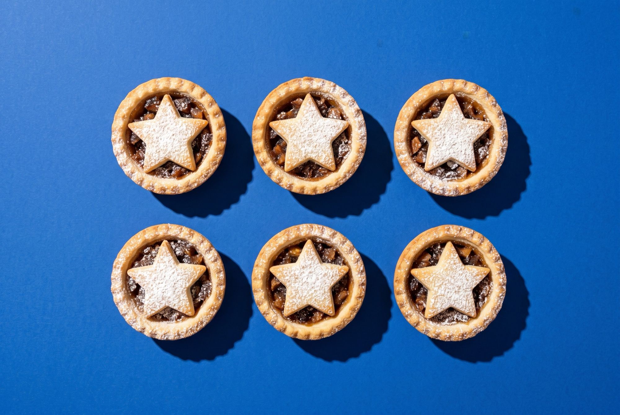 Small festive mince pies with pastry star tops dusted with icing sugar on a cobalt blue backdrop