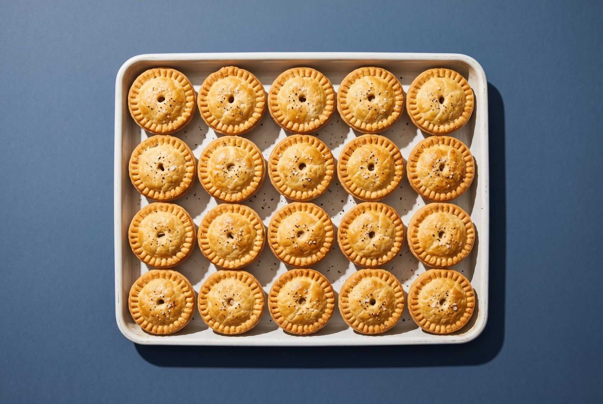 A white tray of twenty mini savoury pies with crimped golden crusts, seen from above on a slate blue backdrop