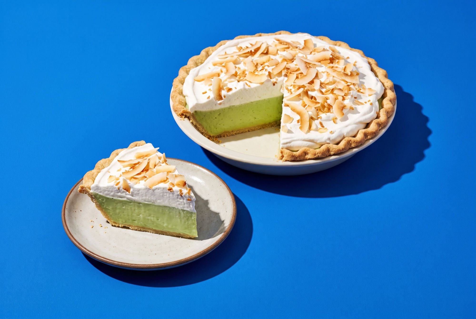 A pandan coconut cream pie with a pale green custard layer under whipped cream and toasted coconut, one slice on a plate beside it, on a cobalt blue backdrop