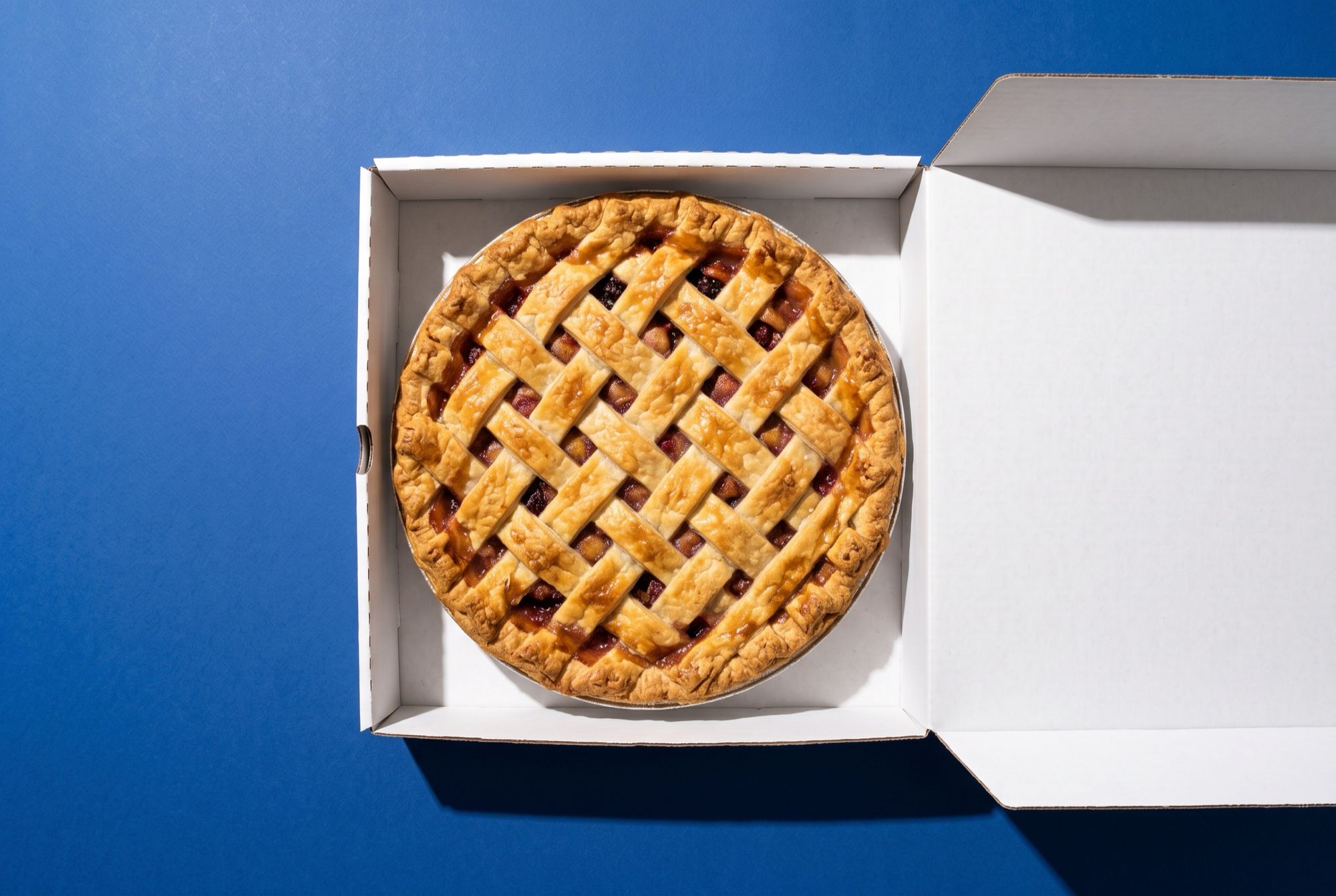 An open plain white pie box holding a whole lattice-top pie, seen from above on a cobalt blue backdrop