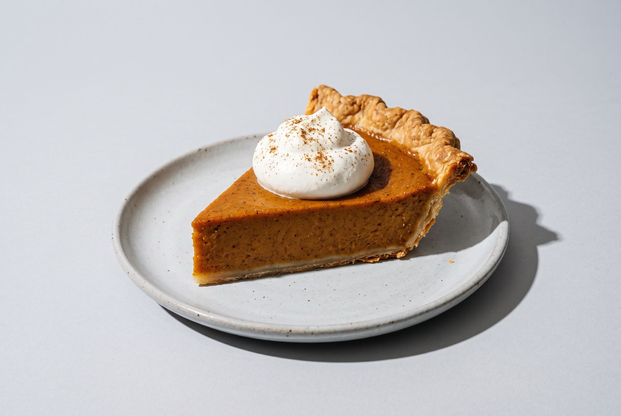A slice of spiced pumpkin pie with a crimped crust edge and a spoonful of whipped cream on a grey plate