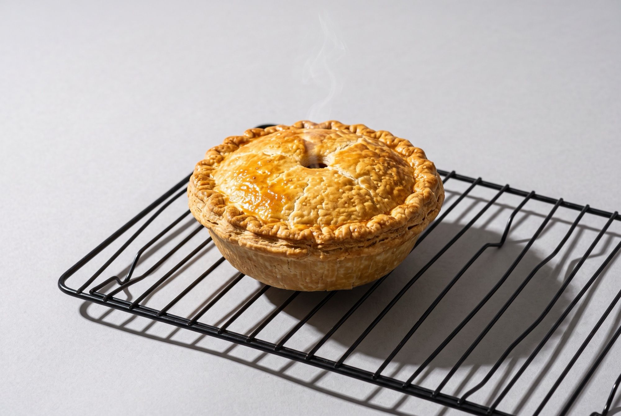 A golden personal pie resting on a black wire cooling rack with a little steam rising